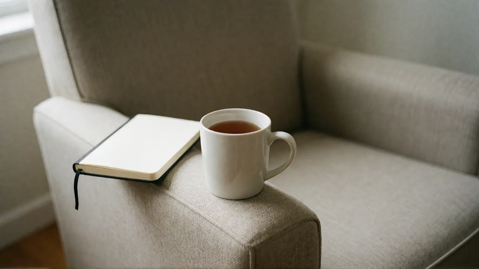 A white mug of tea and a closed blank notebook on the broad arm of an upholstered armchair.