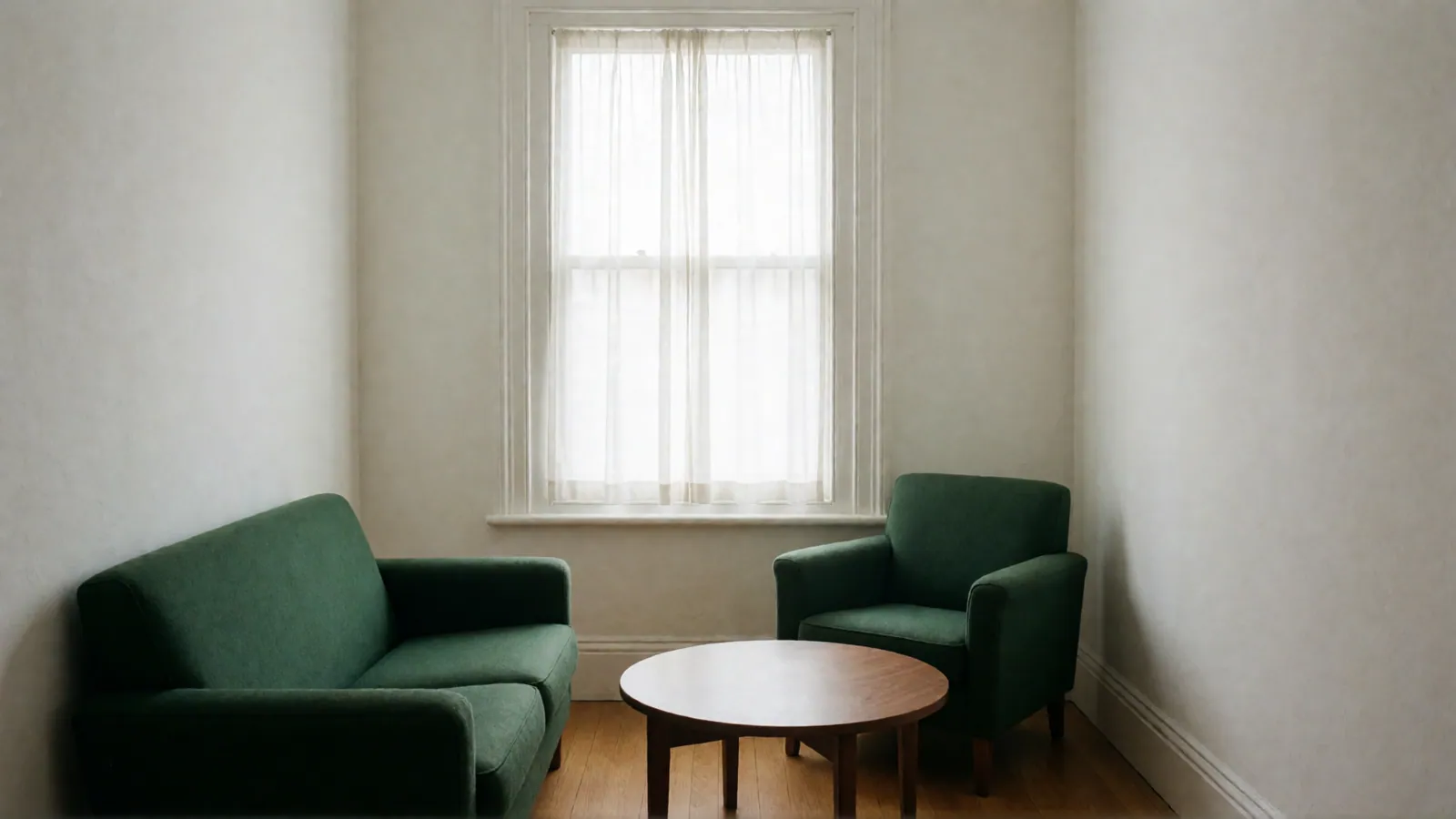 A therapy room with a deep green two-seat sofa and a green armchair around a round walnut table, below a curtained window.