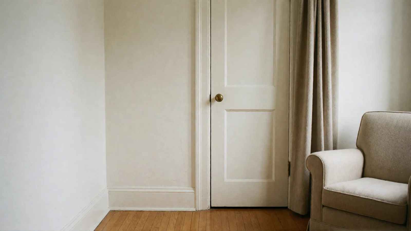 A closed white panel door with a brass knob, a heavy linen curtain beside it and the corner of an oatmeal armchair, in soft light.