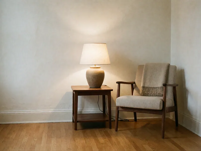A warm table lamp lit on a walnut side table beside a wooden-armed chair with a folded throw, in the corner of a quiet room.