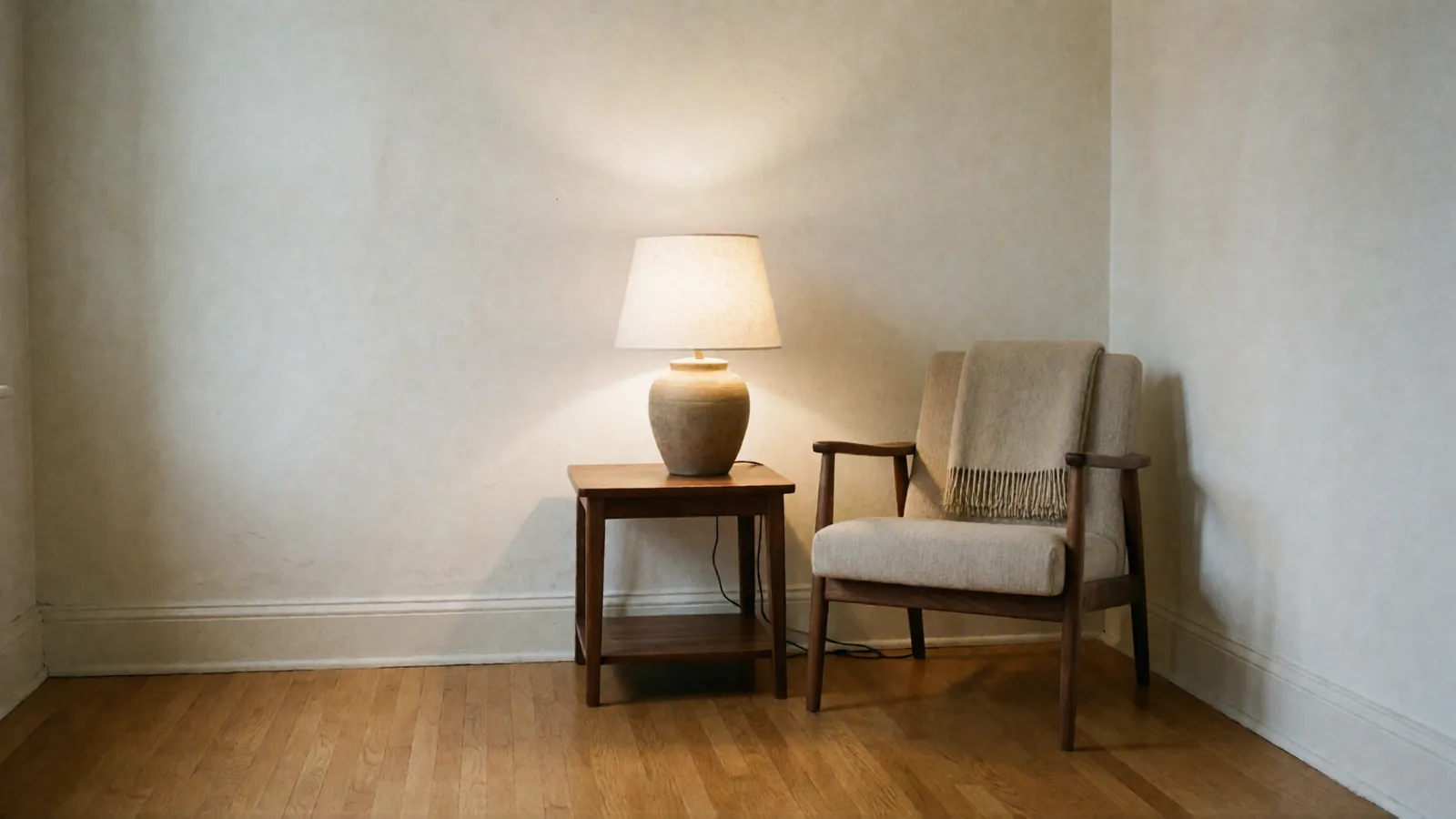 A warm table lamp lit on a walnut side table beside a wooden-armed chair with a folded throw, in the corner of a quiet room.