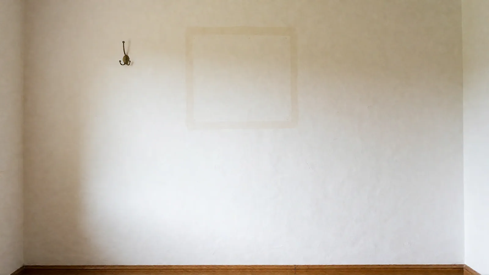 A bare white plaster wall with one empty brass hook and the faint pale square where a frame once hung.