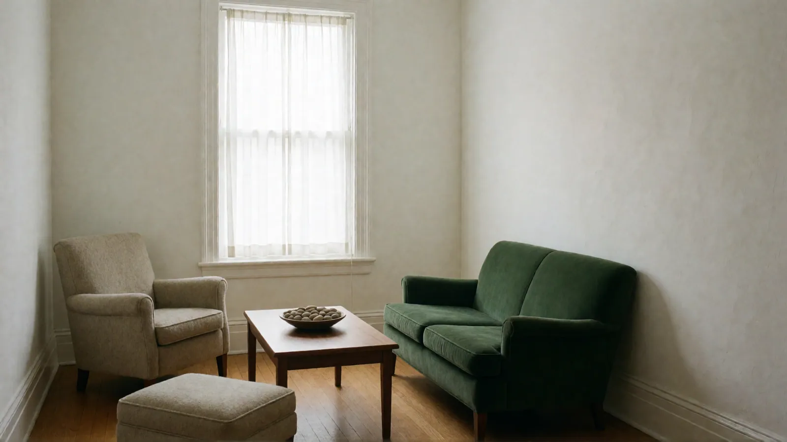 A calm room with a deep green velvet sofa, an oatmeal armchair and an upholstered footstool around a walnut coffee table holding a bowl of smooth stones, below a curtained window.