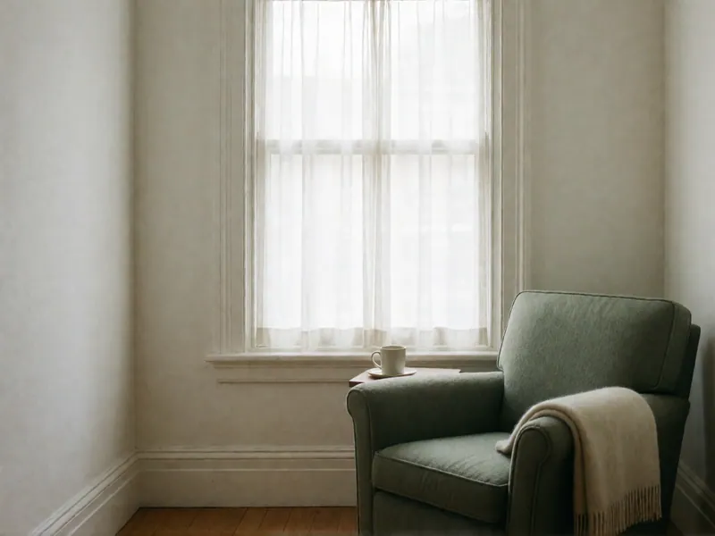 A sage armchair with a cream throw over its arm beside a curtained window, a mug of tea on the sill.