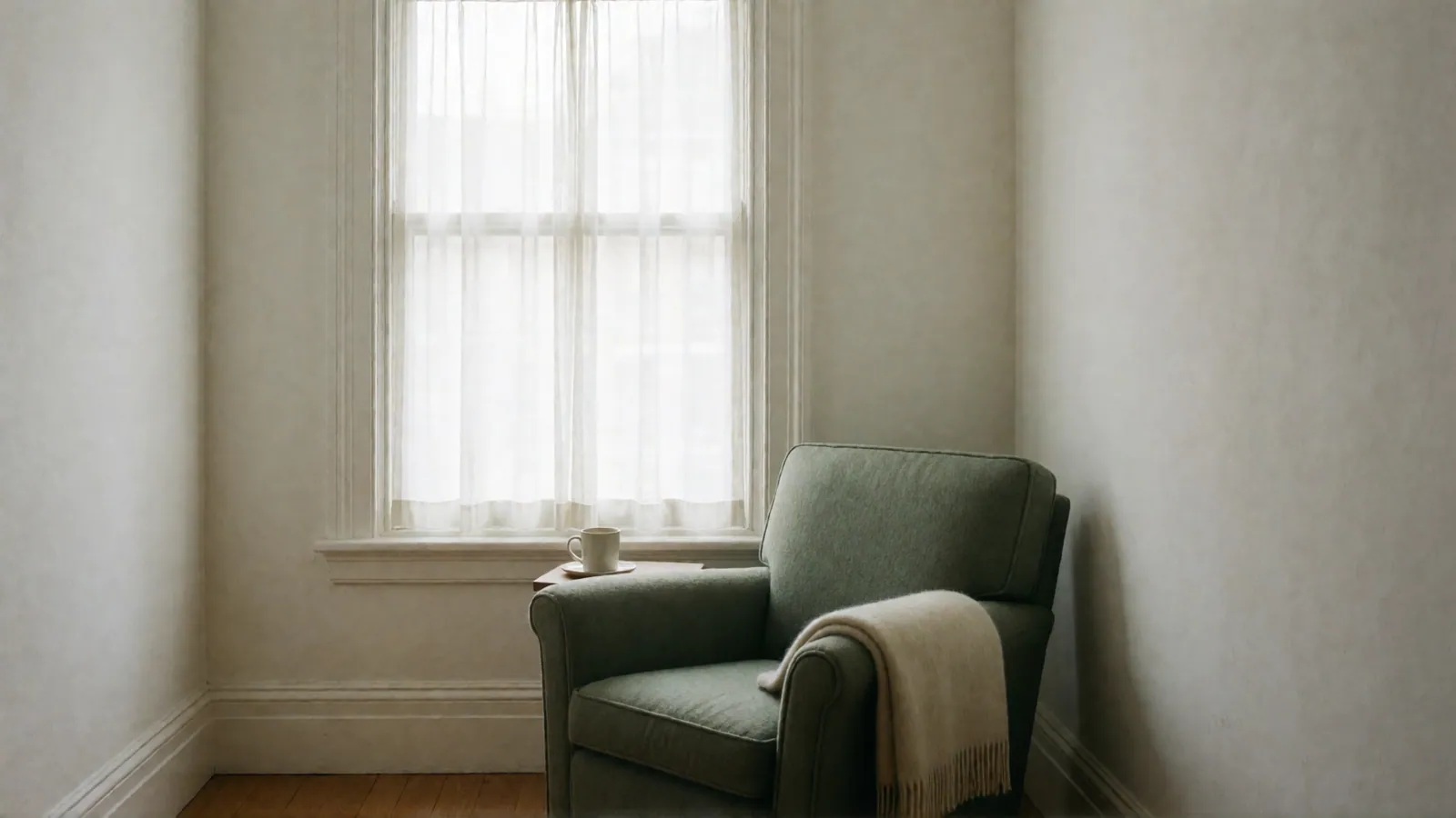A sage armchair with a cream throw over its arm beside a curtained window, a mug of tea on the sill.