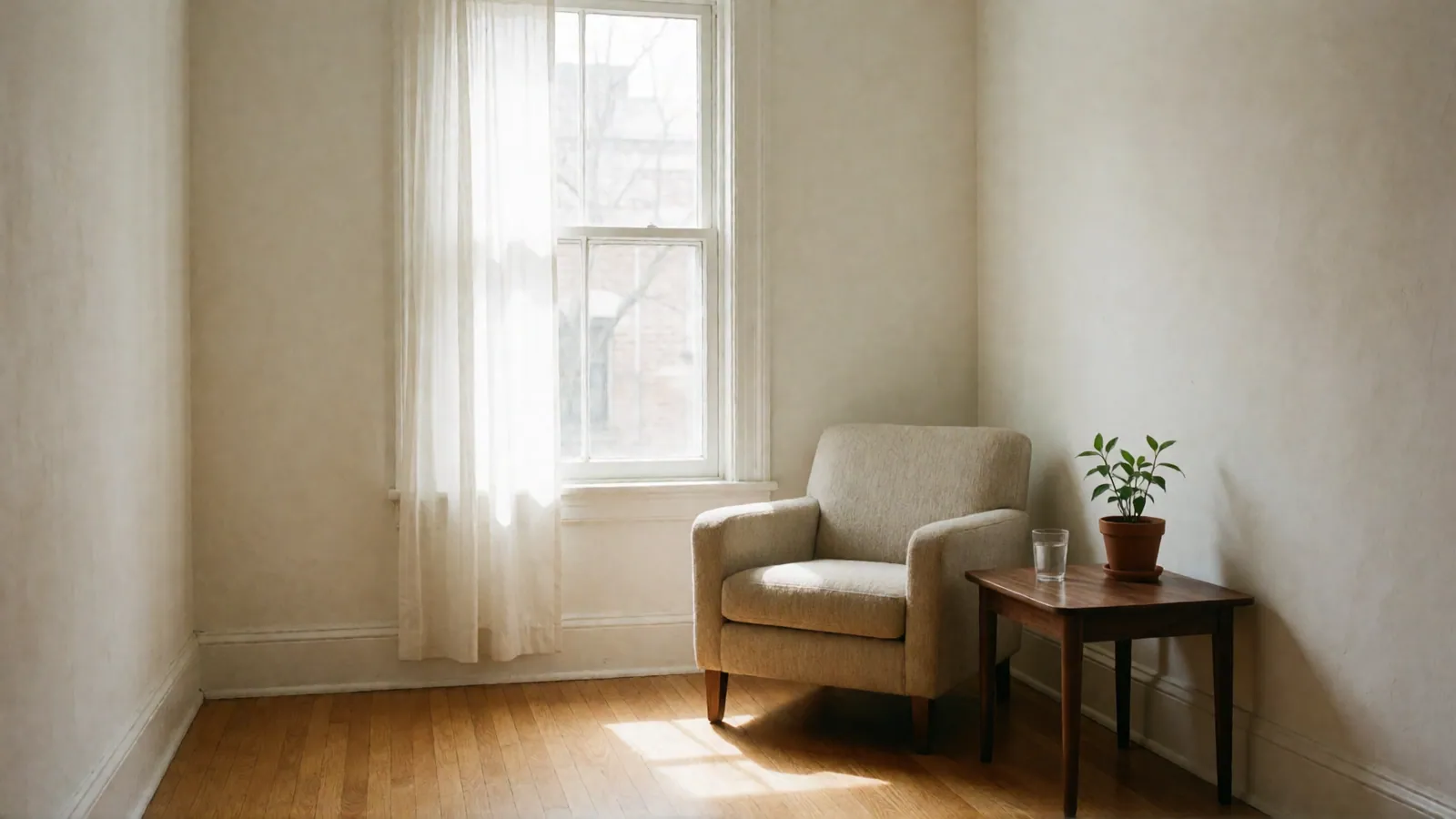 An oatmeal armchair beside a walnut side table with a small plant and a glass of water, sunlight falling across the oak floor.