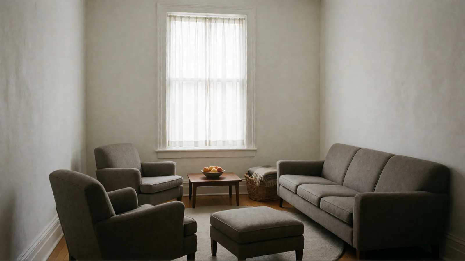A larger room with a long gray sofa, two gray armchairs and an ottoman around a round rug, a low table with a bowl of fruit and a basket of blankets.