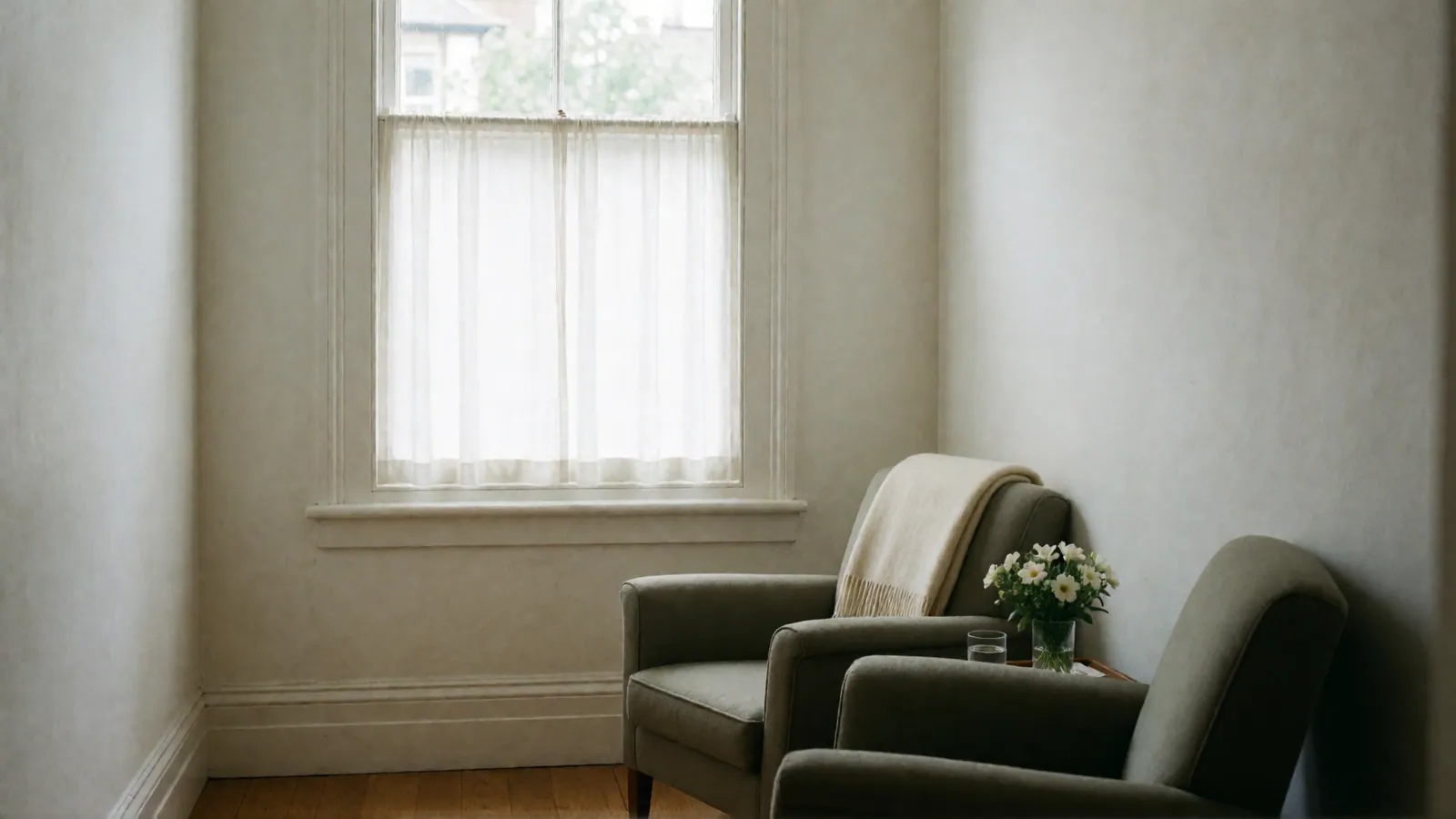 Two gray armchairs side by side at a slight angle, a cream throw on one, a small vase of white flowers and a glass of water between them.