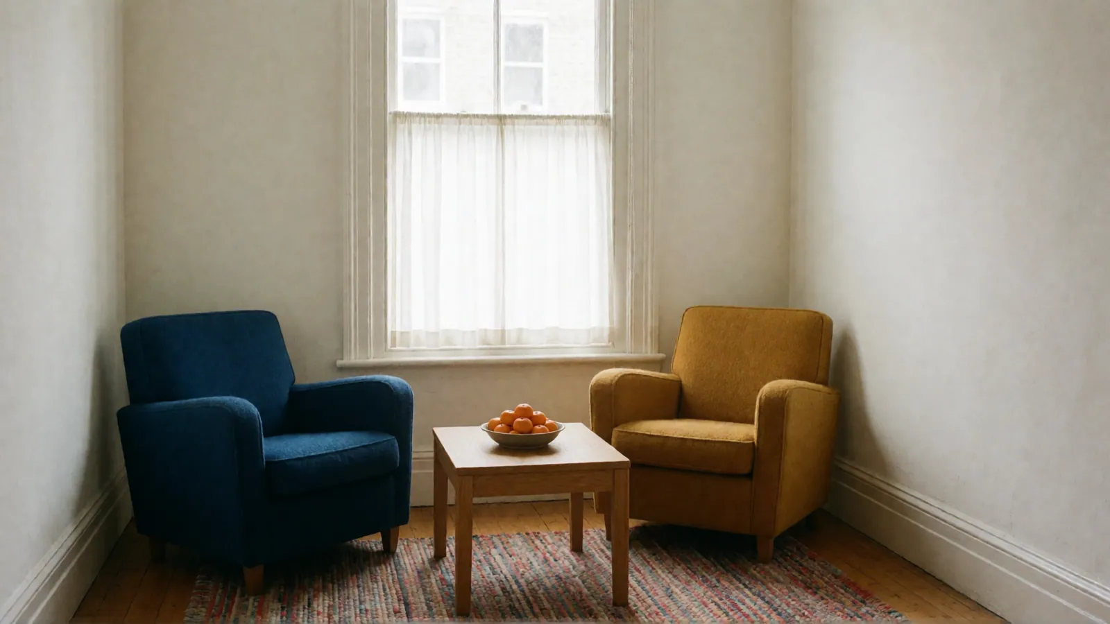 A deep blue armchair and a mustard armchair angled toward each other on a woven rug of soft colors, a low oak table with a bowl of clementines between them.