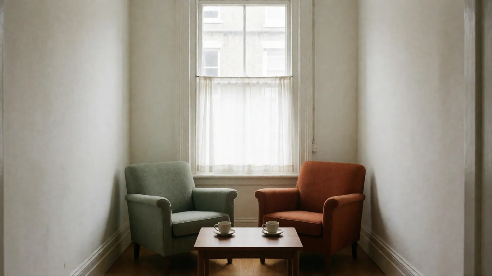A sage armchair and a rust armchair turned slightly toward each other at a low walnut table with two cups of tea, below a window.