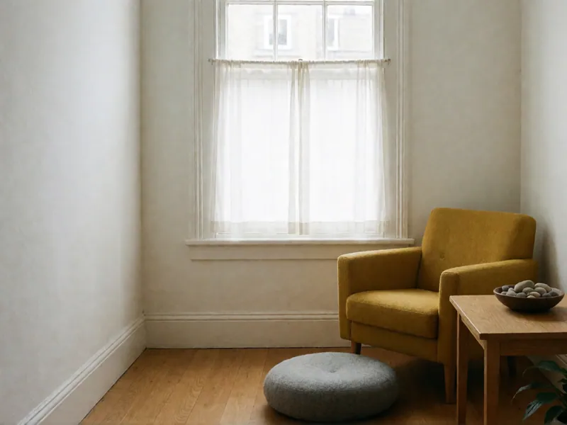A mustard armchair, a round gray felt floor cushion and a small oak table with a bowl of stones and a wooden puzzle cube, below a window.
